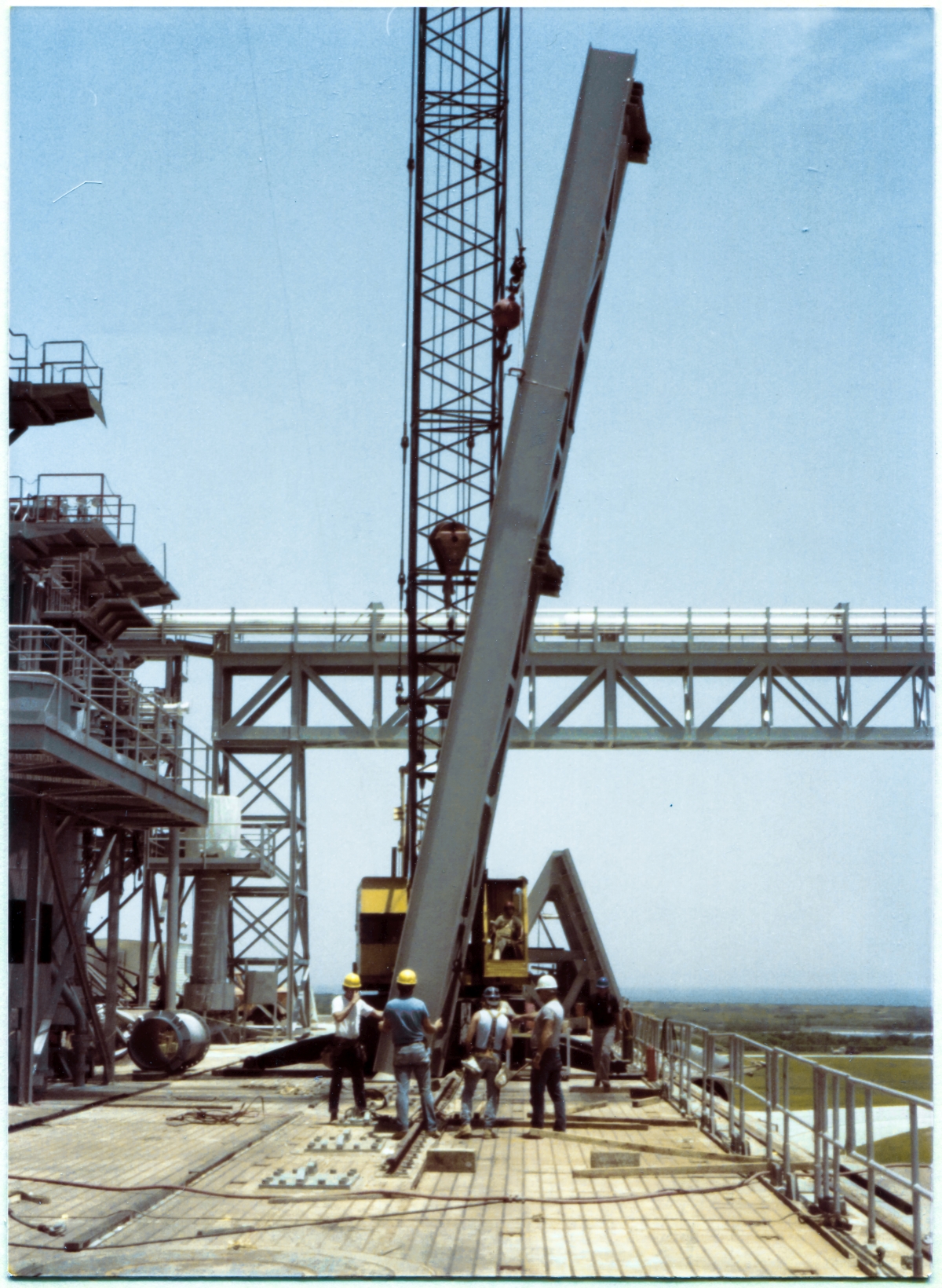 Image 073. The GOX Arm Strongback remains suspended just above the Pad Deck, still within easy reach, as our small group works out the precise arrangements which will need to be effected from here on, as the Strongback rises above the ground, far out of reach, until it arrives at its destination on the Fixed Service Structure, 250 feet straight up from the Pad Deck it now hangs less than a yard above. For the moment, it is rotated 180 degrees away from its final orientation, flush against the Primary Framing on side 1 of the FSS, extending from elevation 260'-0” to 300'-0”. During lifts, orientations of that which is being lifted do not necessarily match their final orientations, and this can be for any number of reasons, most of which are only apparent to those persons directly involved in the lift, and none of which might be visually apparent to an untrained or uninvolved observer. Photo by James MacLaren.